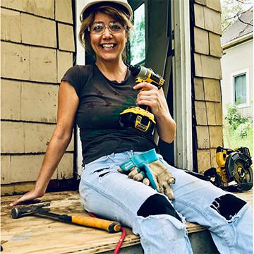 Young woman in a hard hat holding a drill sitting on a porch of a house for Habitat for Humanity